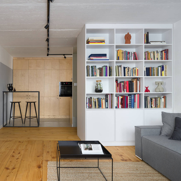 Stylish living room with pine wood floor and kitchen area behind white bookcase - 写真・画像