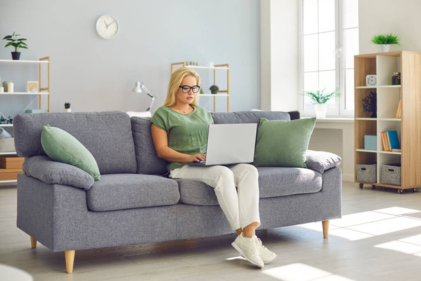 Young woman in eyeglasses sitting on couch and working on project using laptop computer - Photo, image