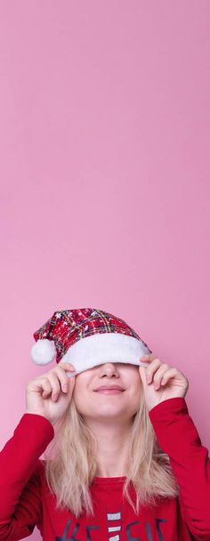 ピンクの背景に隔離された赤いサンタクラスの帽子の美しい女性の肖像画。美しい女の子幸せと興奮している。楽しいクリスマスと新年の休日。パノラマ。コピースペース。旗 - 写真・画像