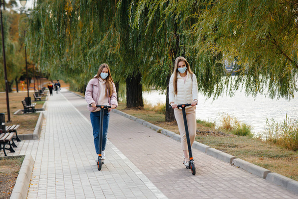 Two young beautiful girls in masks ride electric scooters in the Park on a warm autumn day. Walk in the Park. - Valokuva, kuva