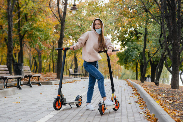 Młoda piękna dziewczyna w masce jedzie w parku na skuterze elektrycznym w ciepły jesienny dzień. Spacer po parku. - Zdjęcie, obraz