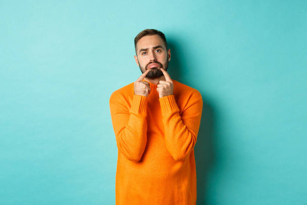 Imagem de homem barbudo sombrio, fazendo cara triste e franzina, de pé chateado em suéter laranja contra fundo turquesa - Foto, Imagem