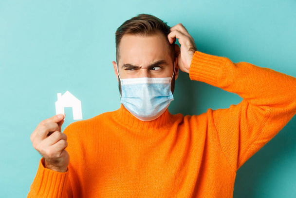 Emlak ve koronavirüs salgını konsepti. Yüzü maske takmış kafası karışmış bir adam küçük kağıt eve bakarken açık mavi stüdyo arka planında turuncu süveter giyiyor. - Fotoğraf, Görsel