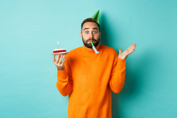 Happy birthday guy celebrating, wearing party hat, blowing wistle and holding bday cake, standing against white background - Fotoğraf, Görsel