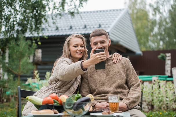 Modern husband and wife have fun taking an emotional selfie together during a picnic. Sitting at a table with vegetables, barbecue meat and beer. - Фото, изображение