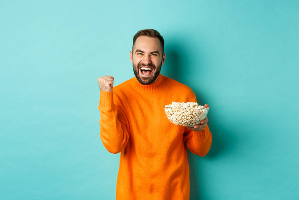 Hombre guapo alegre en suéter naranja, diciendo sí, animando y regocijándose, comiendo palomitas de maíz y viendo deportes, haciendo bomba de puño satisfecho, de pie sobre fondo azul - Foto, Imagen