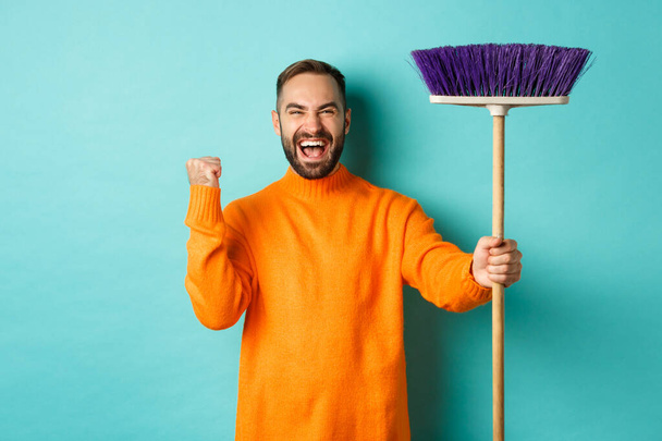 Образ воодушевленного и мотивированного человека, готовящегося к уборке, держащего метлу и делающего жест кулачного насоса, стоящего на бирюзовом фоне - Фото, изображение