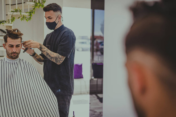 Młody mężczyzna z wytatuowanymi ramionami obcina facetowi włosy w salonie fryzjerskim - Zdjęcie, obraz