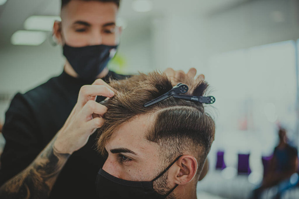 Молодой человек с татуированными руками стрижет мужчине волосы в парикмахерской - Фото, изображение