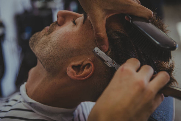 detail mladého muže s vytetovanými pažemi stříhá mužské vlasy v holičství - Fotografie, Obrázek