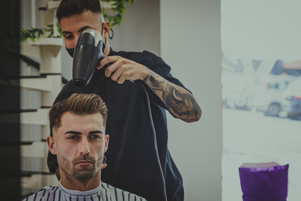 um jovem com braços tatuados corta o cabelo de um homem em uma barbearia - Foto, Imagem
