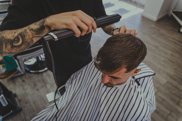 un giovane uomo con le braccia tatuate taglia i capelli di un uomo in un barbiere - Foto, immagini