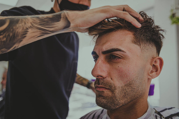 nuori mies, jolla on tatuoidut kädet, leikkaa miehen hiukset parturissa - Valokuva, kuva