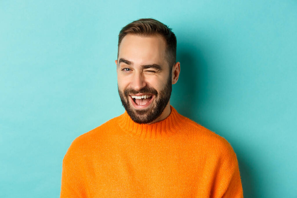 Όμορφος άνθρωπος με αυτοπεποίθηση να κλείνει το μάτι στην κάμερα, χαμογελαστός αυθάδης, στέκεται σε πορτοκαλί πουλόβερ με φόντο το μπλε - Φωτογραφία, εικόνα