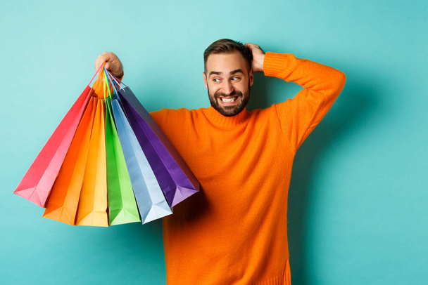 Handsome young man looking awkward and scratching head, staring at shopping bags with gifts, standing over turquoise background - Photo, Image