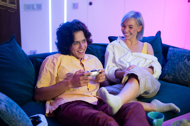 Νέοι χαρούμενοι Καυκάσιοι σύζυγοι παίζουν βιντεοπαιχνίδια ο ένας εναντίον του άλλου στο σπίτι απολαμβάνοντας τον ελεύθερο χρόνο τους σε αργή κίνηση - Φωτογραφία, εικόνα