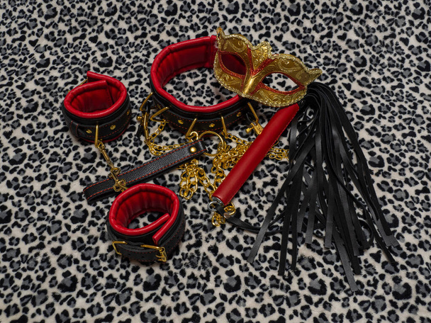 Toys for adults. Carnival mask, red and black leather handcuffs, collar, whip and nipple clamps with a gold chain on a leopard background. BDSM equipment. - Photo, Image