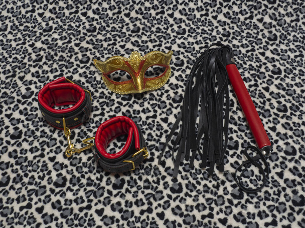 Toys for adults. Carnival mask, red and black leather handcuffs and a whip on a leopard background. BDSM equipment. - Photo, Image