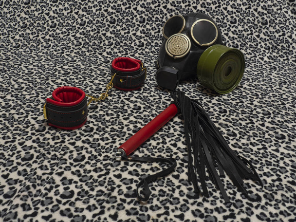 Παιχνίδια για ενήλικες. Μάσκα αερίου, κόκκινες και μαύρες δερμάτινες χειροπέδες και ένα μαστίγιο σε φόντο λεοπάρδαλης. Εξοπλισμός BDSM. - Φωτογραφία, εικόνα