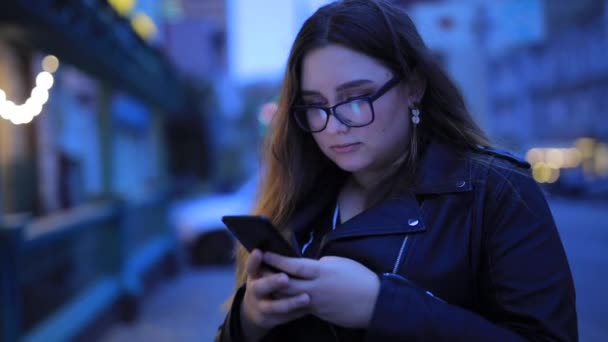 Nainen chattailee älypuhelimessa iltakaupungin taustaa vasten - Materiaali, video