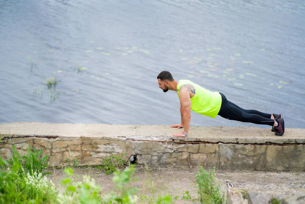 Sporty guy doing push ups or press ups exercise for abs muscles, standing in plank pose, working out outdoor near water - Photo, Image