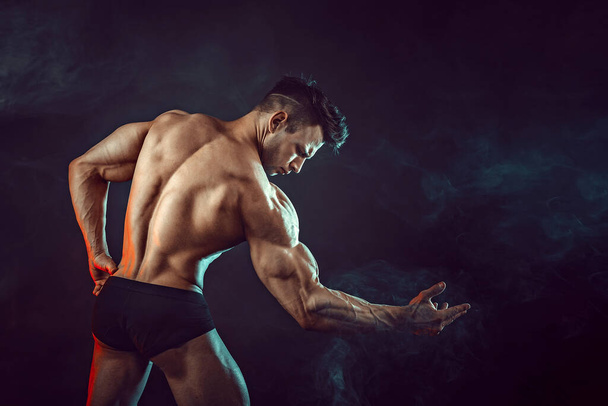Athletic man flexing muscles in studio on dark background with smoke. Strong bodybuilder with perfect abs - Photo, image