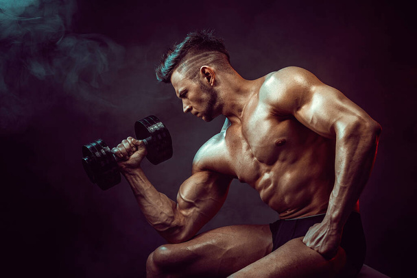 Athletic man flexing muscles in studio on dark background with smoke. Strong bodybuilder with perfect abs - Фото, зображення