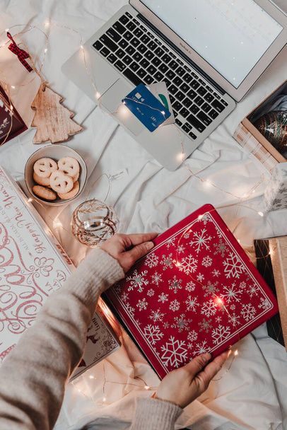 Christmas holiday online shopping. Gift boxes, lights. Festive season. Using phone and credit card. Woman in cozy sweater and jeans uses laptop for shopping. Flat lay. Christmas boxes, ornaments. - Фото, изображение