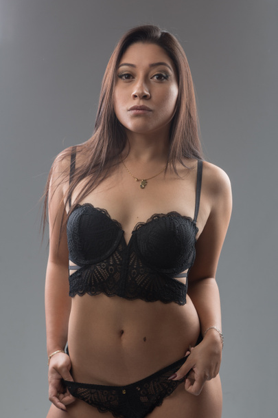 Young slim latin girl with black hair, wears makeup, jewelry and black lace lingerie, studio with gray background - Φωτογραφία, εικόνα