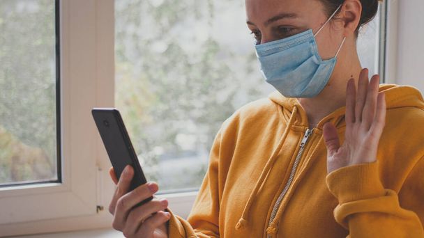 Young woman in medical protective mask making video call with smartphone, gesturing hi to friend or parent, standing near window indoor on self isolation. Social media, stay at home at quarantine - Photo, image