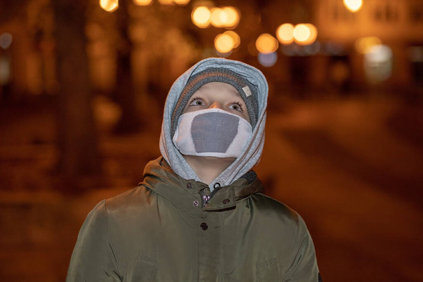 Nahaufnahme Porträt eines kaukasischen Teenagers in Maske und warmer Freizeitkleidung, der auf der Straße steht und nach oben schaut. Kleiner männlicher Teenager im Nachtlicht der Stadt. - Foto, Bild