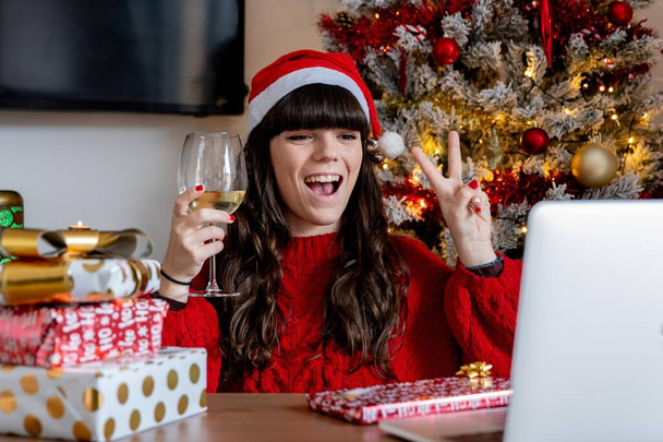 Νεαρό κορίτσι με το χριστουγεννιάτικο καπέλο και το κόκκινο πουλόβερ κάνει μια βιντεοκλήση στην οικογένειά της για να τους δείξει τα δώρα Χριστουγέννων, ενώ πίνοντας ένα ποτήρι κρασί με το χριστουγεννιάτικο δέντρο πίσω - Φωτογραφία, εικόνα