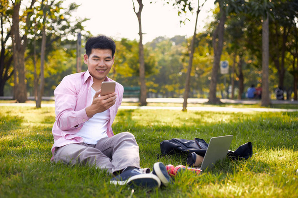 Ασιάτης ελεύθερος επαγγελματίας κάθεται σε ένα πάρκο χρησιμοποιώντας το τηλέφωνό του σε μια άνετη εργάσιμη ημέρα. - Φωτογραφία, εικόνα