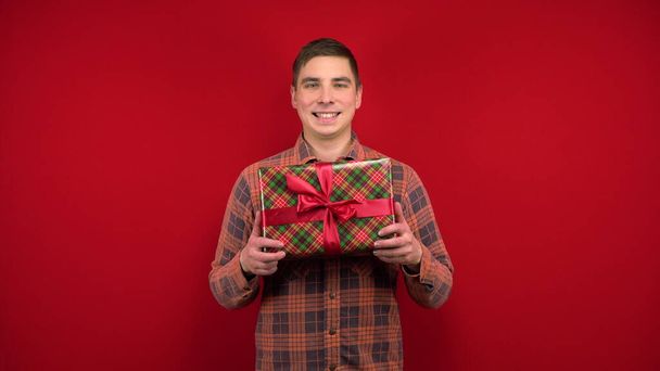 Młody mężczyzna trzyma prezent świąteczny i raduje się. Kręcenie w studio na czerwonym tle. - Zdjęcie, obraz
