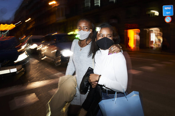 Meşgul bir şehirde gece vakti alışveriş torbalarıyla yürüyen iki Afro-Amerikan kadının flaşlı portresi. Şehir ışıkları ve hareket bulanıklığı - Fotoğraf, Görsel