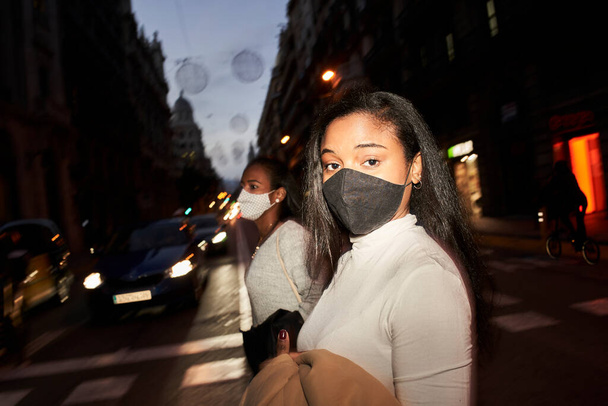 Fotografia di strada di due donne afroamericane in maschera che attraversano la strada di notte in una città trafficata. Uno di loro guarda seriamente la telecamera. Luci di città e movimento sfocato. Lunga esposizione - Foto, immagini