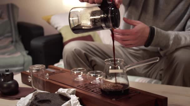 Un hombre desconocido vierte té preparado de una tetera en una jarra, de una jarra en un tazón de vidrio y bebe mientras está sentado en un sofá - Metraje, vídeo
