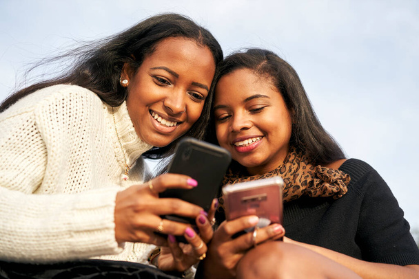 Nahaufnahme zweier schwarzer Mädchen auf einer Treppe in einer Stadt, während sie etwas Interessantes auf ihrem Handy beobachten. Mensch und Technik-Konzept mit Smartphone. - Foto, Bild
