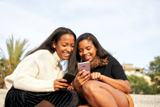 Две улыбающиеся черные девушки на ступеньках в городе и с помощью мобильного телефона. Концепция людей и технологий со смартфоном. - Фото, изображение