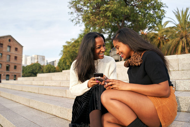 Twee zwarte meisjes op een trap in een stad terwijl ze een mobiele telefoon gebruiken en lachen. Mensen en technologie concept met smartphone. - Foto, afbeelding