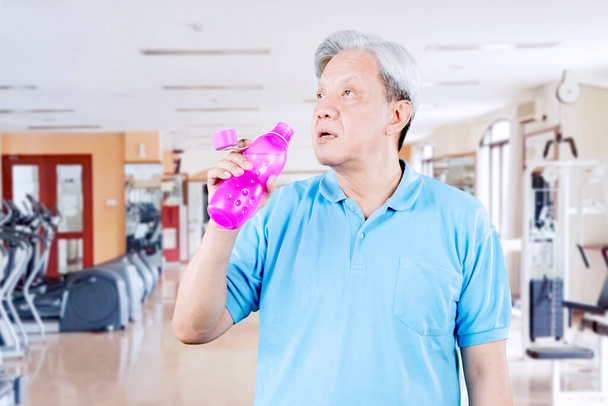 Senior man wearing sportswear while drinking a bottle of water after exercise in the fitness center - Фото, зображення
