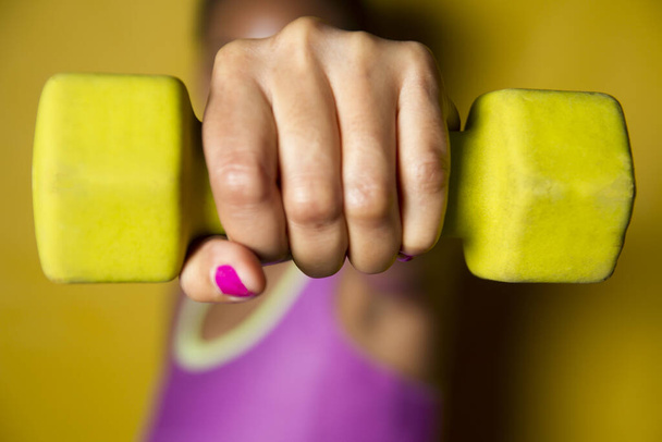 selective focus of one unrecognizable young woman fist lifting dumbbell weight exercising lifting dumbbells on yellow background - Photo, Image