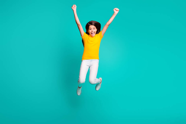 Full length photo of ecstatic girl jump raise fists wear yellow t-shirt white pants trousers isolated on turquoise color background - 写真・画像