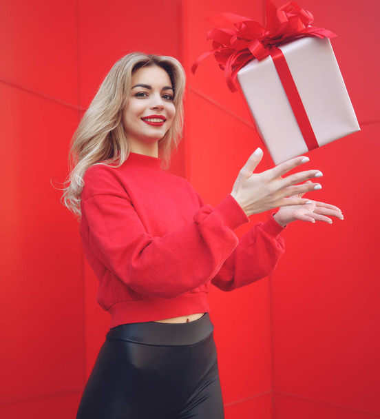 Mujer rubia feliz en la celebración de caja de regalo y mirando a la cámara mientras disfruta sobre fondo rojo. - Foto, imagen
