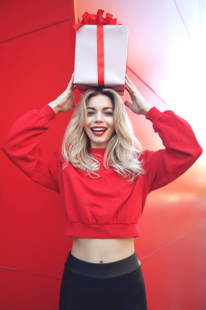 Happy blonde woman in holding gift box and looking at camera while enjoys over red background. - Φωτογραφία, εικόνα