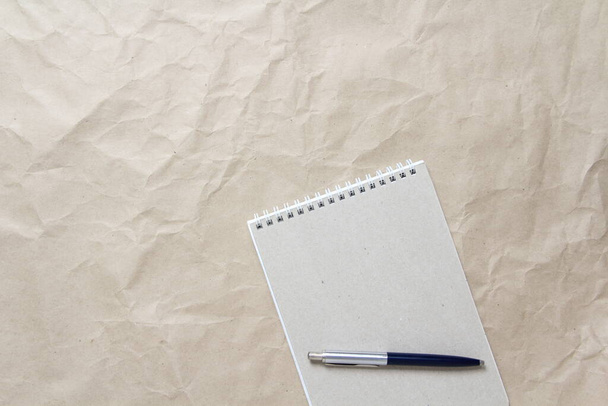 Gray notepad with white coiled spring and pen on a background of beige crumpled craft paper - Foto, afbeelding