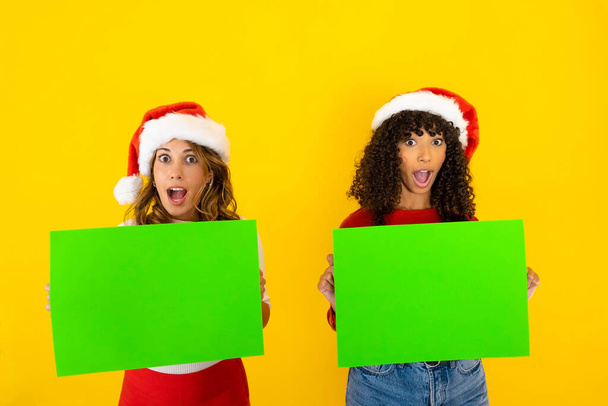 Οριζόντια αστεία χριστουγεννιάτικο πανό με δύο μικτή φυλή γυναικών με το στόμα και τα μάτια ορθάνοιχτα στην έκπληκτη έκφραση κρατώντας πράσινο καλάθι για αντίγραφο χώρου ADV μήνυμα σε chroma κλειδί σε κίτρινο φόντο - Φωτογραφία, εικόνα