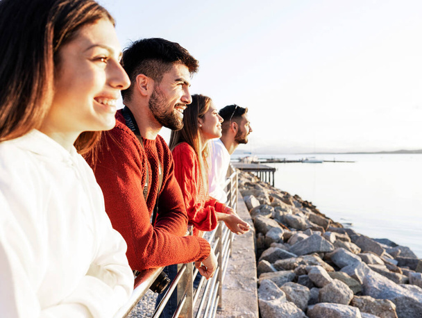 Young beautiful happy people leaning on a metal railing at the sunset on the water in sea winter vacation resort - Confident friends group looking at the horizon - Focus on closer bearded man - Foto, Imagen