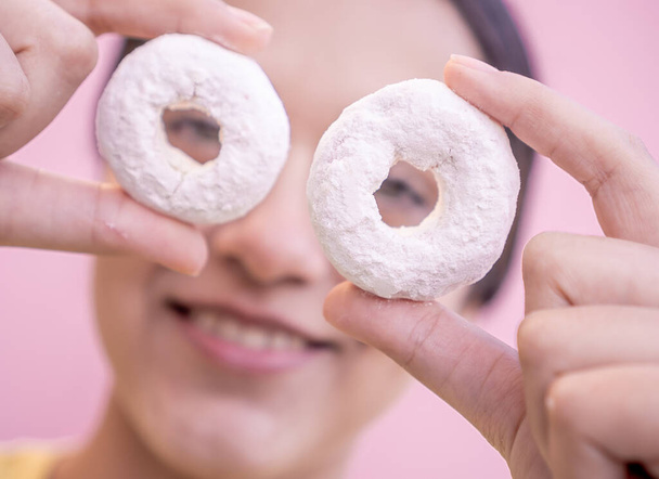 Λατίνα γυναίκα κρατώντας δύο νόστιμα ντόνατς πασπαλισμένα με ζάχαρη γυαλί μπροστά από τα μάτια της με ένα ροζ φόντο σε στενή έννοια. - Φωτογραφία, εικόνα