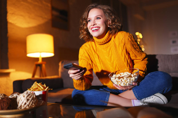 Vidám nő egy tál friss popcornnal, mosolyog és csatornát vált a tévében, miközben otthon ül a kanapén. - Fotó, kép